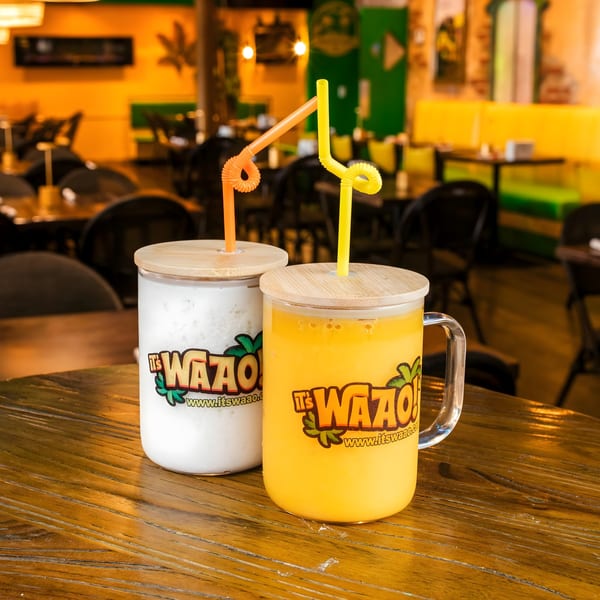 Two "It's Waao!" branded drinks, one white and one yellow, with straws on a wooden table.