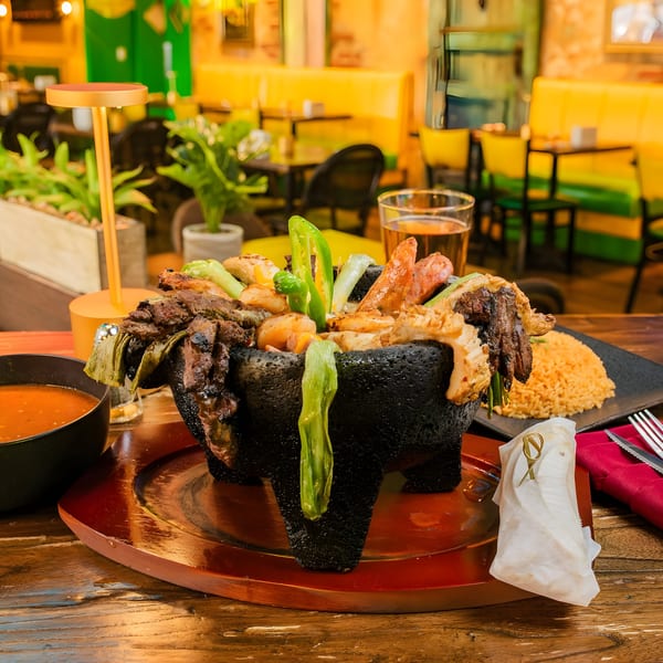Molcajete with grilled meats, shrimp, and peppers, served with rice, soup, and beer in a restaurant.