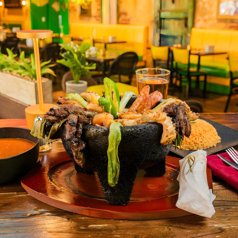 Molcajete with grilled meats, shrimp, and peppers, served with rice, soup, and beer in a restaurant.
