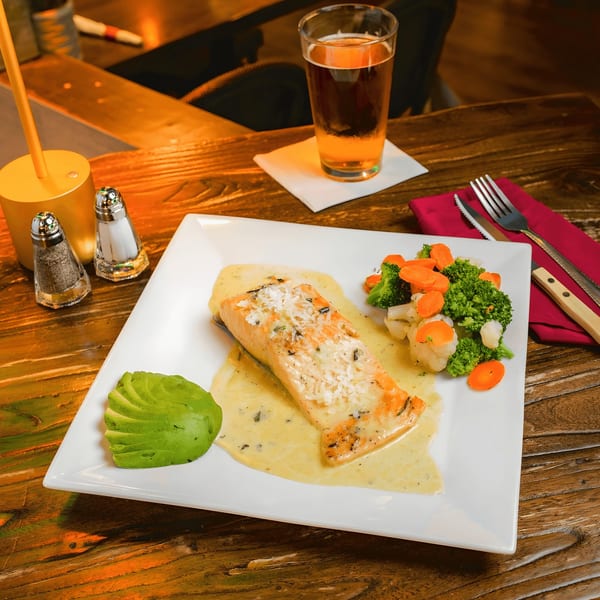 Salmon with cream sauce, sliced avocado, and mixed vegetables on a white plate with a beer.