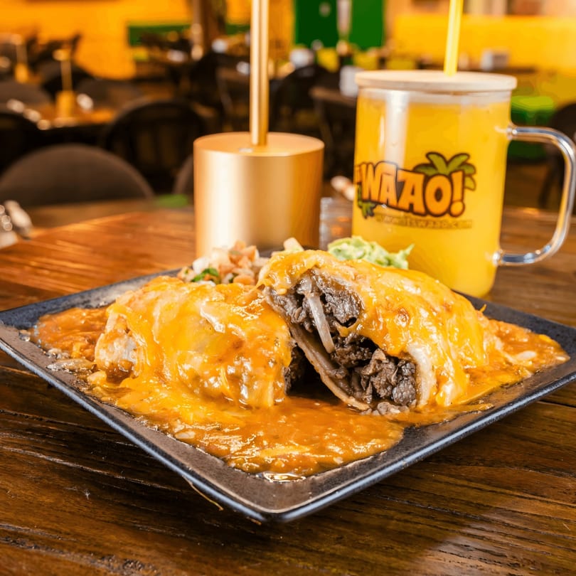 A beef burrito covered in melted cheese and sauce, with a WaZao! branded drink in the background.