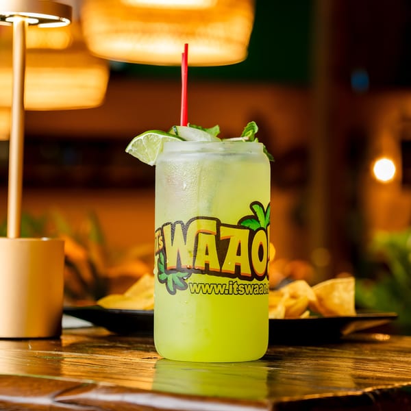A refreshing green drink in a branded glass with lime, mint, and a red straw on a wooden table.